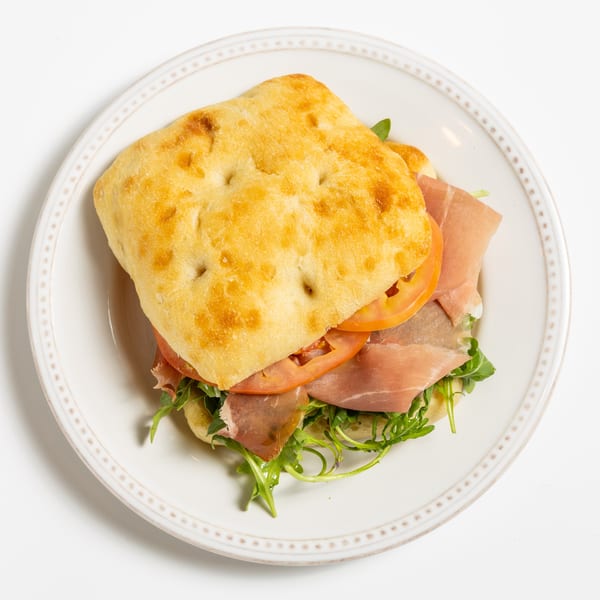 Focaccia sandwich with prosciutto, sliced tomatoes, and arugula on a white plate.