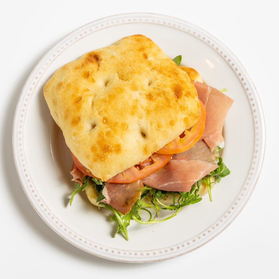 Focaccia sandwich with prosciutto, sliced tomatoes, and arugula on a white plate.