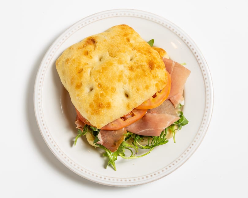 Focaccia sandwich with prosciutto, sliced tomatoes, and arugula on a white plate.