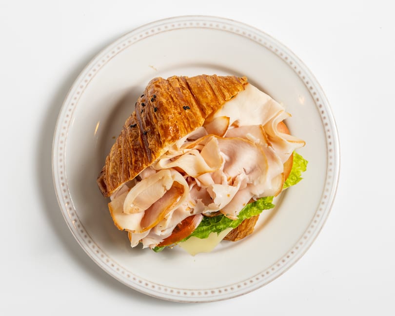 Overhead view of a croissant sandwich with turkey, lettuce, cheese, and tomato on a white plate.