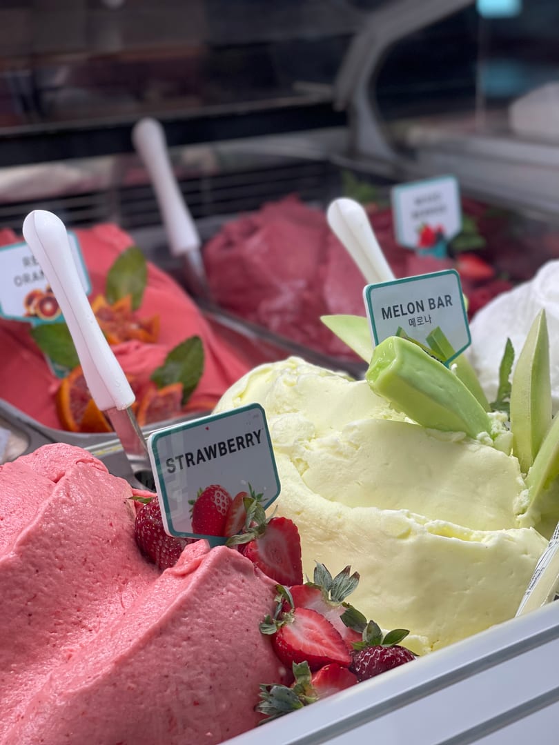 Gelato case with Strawberry, Melon Bar, and Blood Orange flavors garnished with fresh fruit and labeled markers.