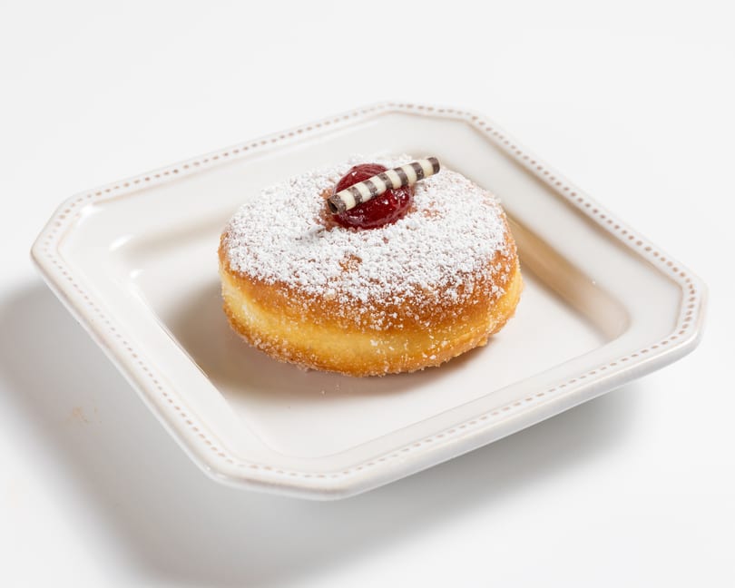 Powdered sugar donut with a dollop of red jam and a striped chocolate curl on a white plate.