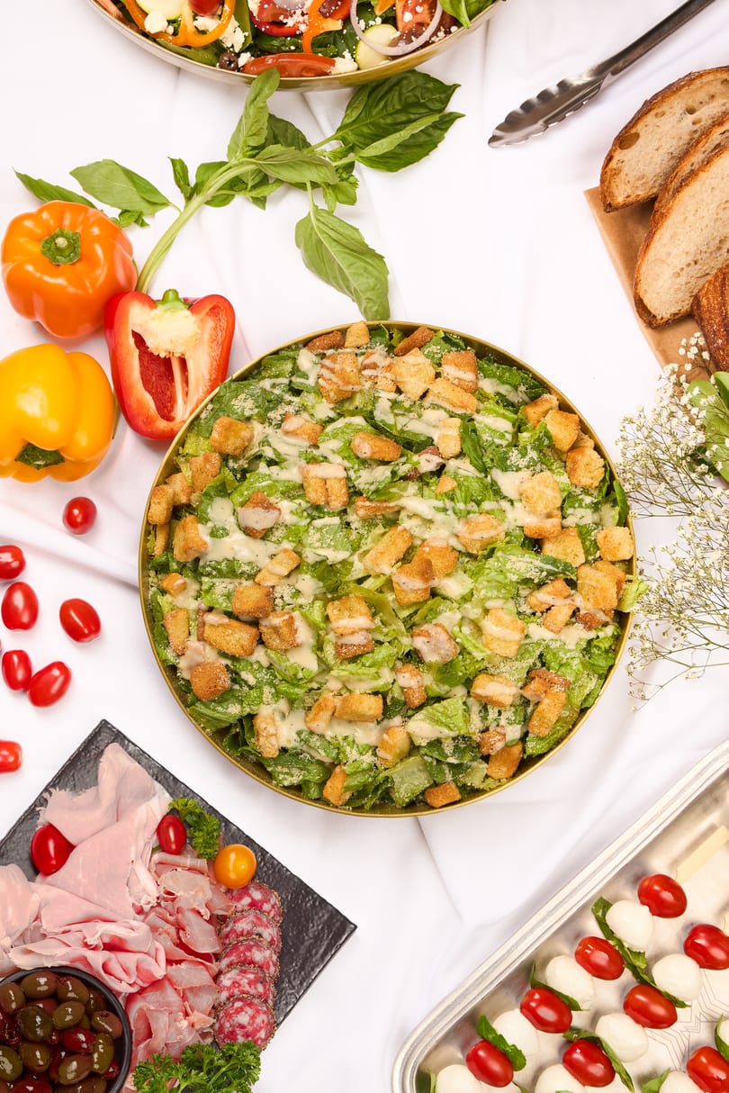 Overhead view of a buffet spread with Caesar salad, Greek salad, charcuterie, and caprese skewers.