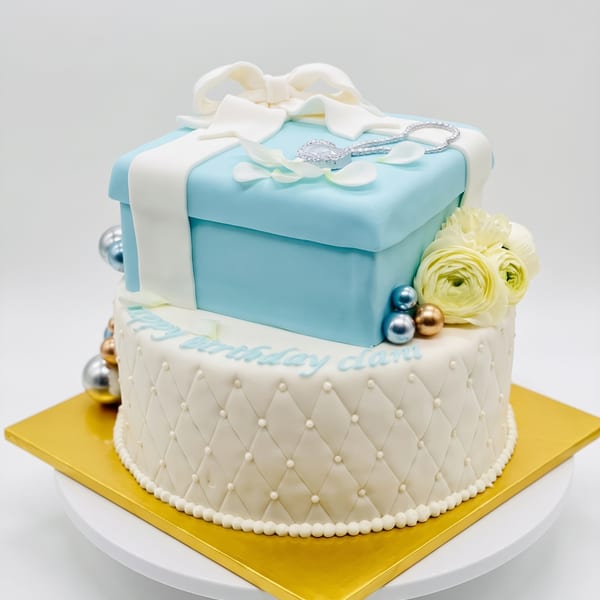 A Tiffany-blue gift box cake with a white fondant bow, jewelry detail, and pearls atop a white quilted tier reading 'Happy Birthday'.
