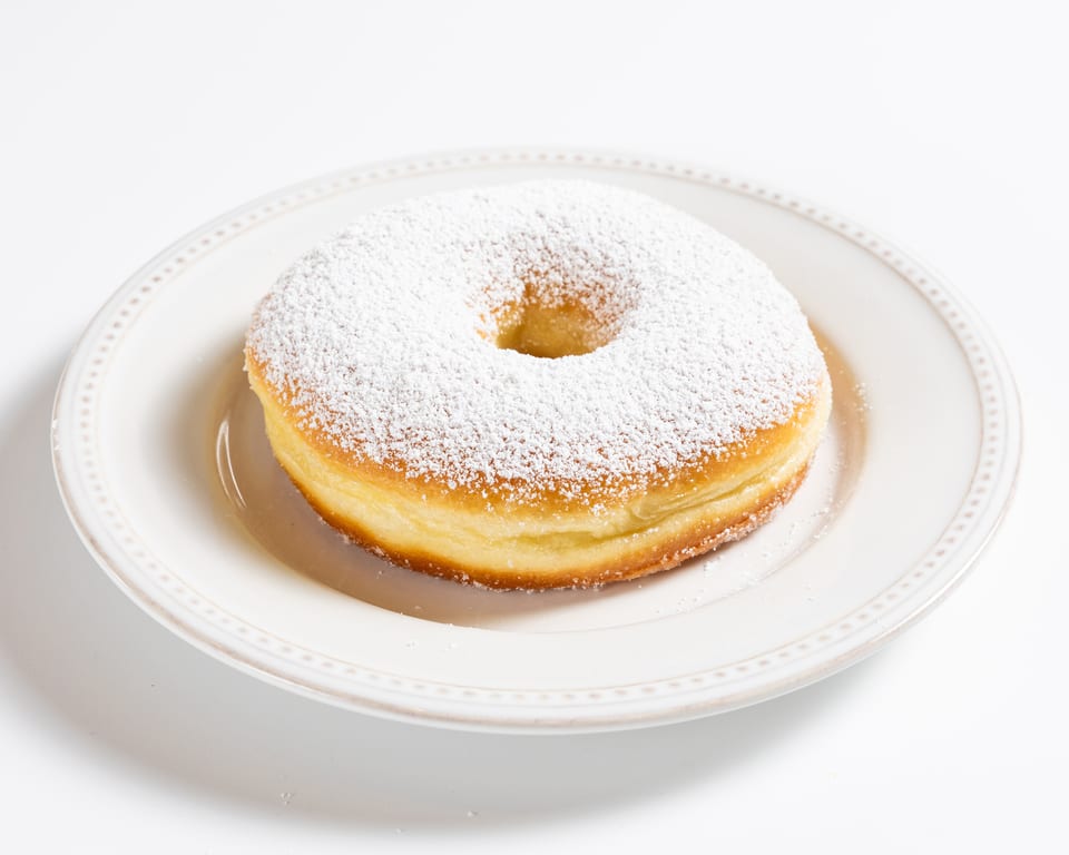 A golden-brown powdered sugar donut on a white plate, against a white background.