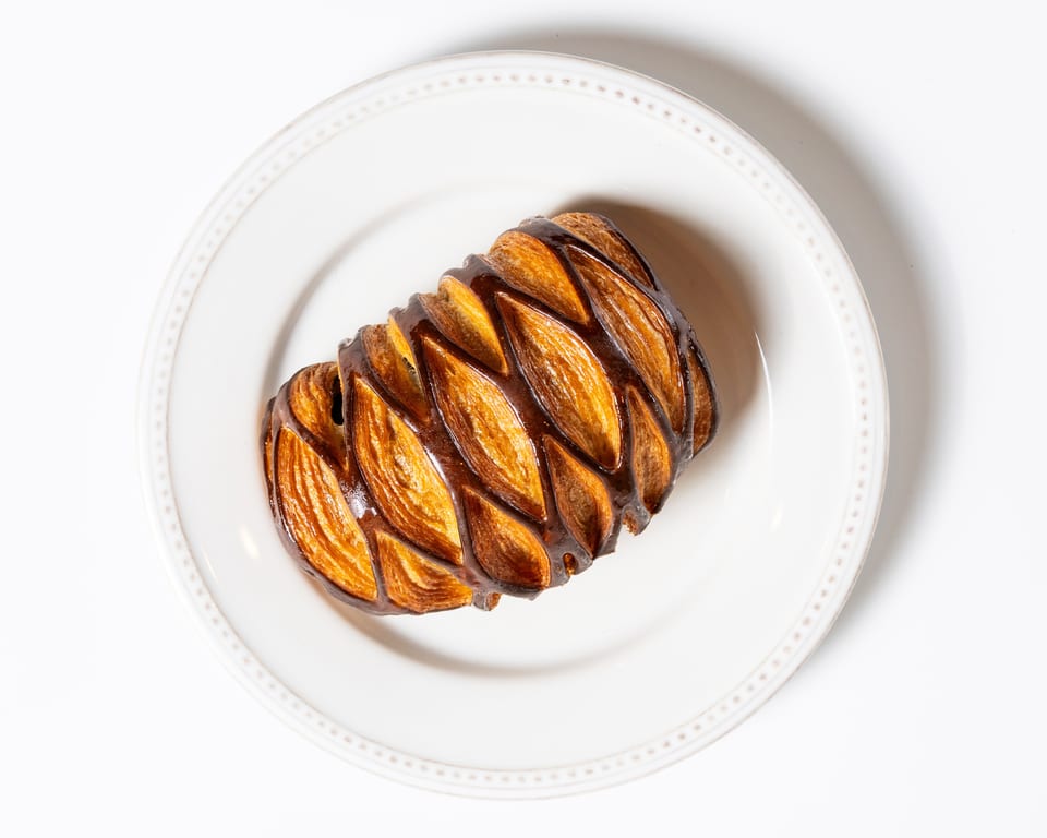 A golden-brown braided pastry with a glossy chocolate drizzle sits on a white plate.