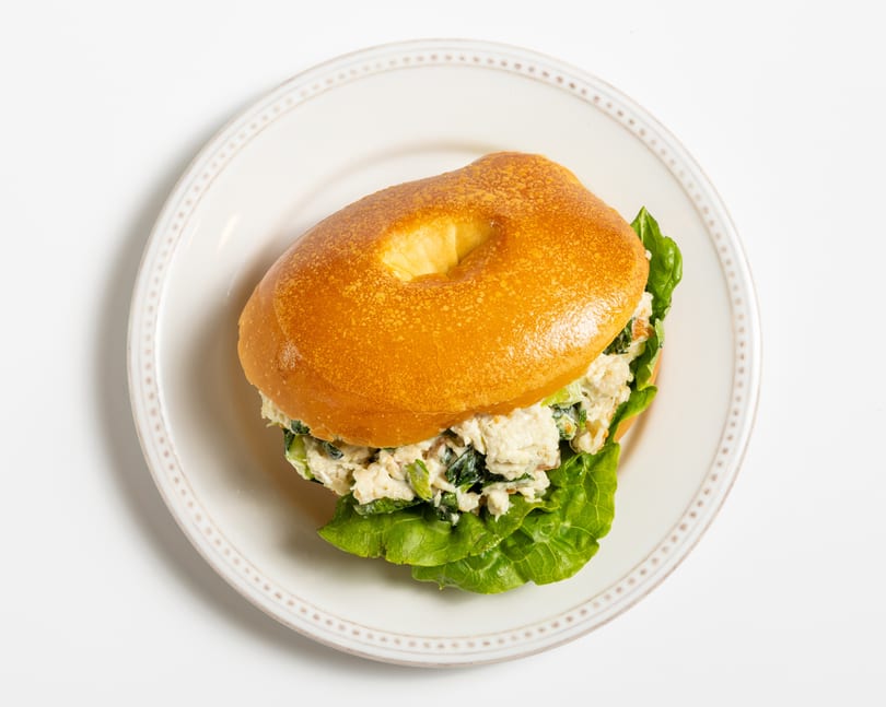 Bagel sandwich with chicken salad and lettuce on a white plate with a decorative rim.