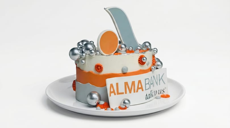 A decorative cake in orange, white, and silver with "ALMA BANK talk to us" sign.
