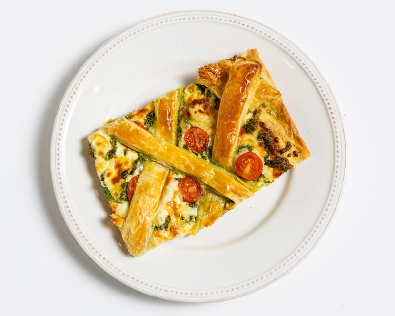 Savory pastry tart with pesto, melted cheese, and cherry tomatoes on a white plate.