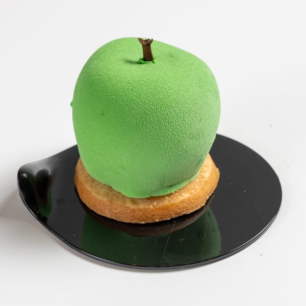 Green apple-shaped dessert with a brown stem on a round cookie and black plate.