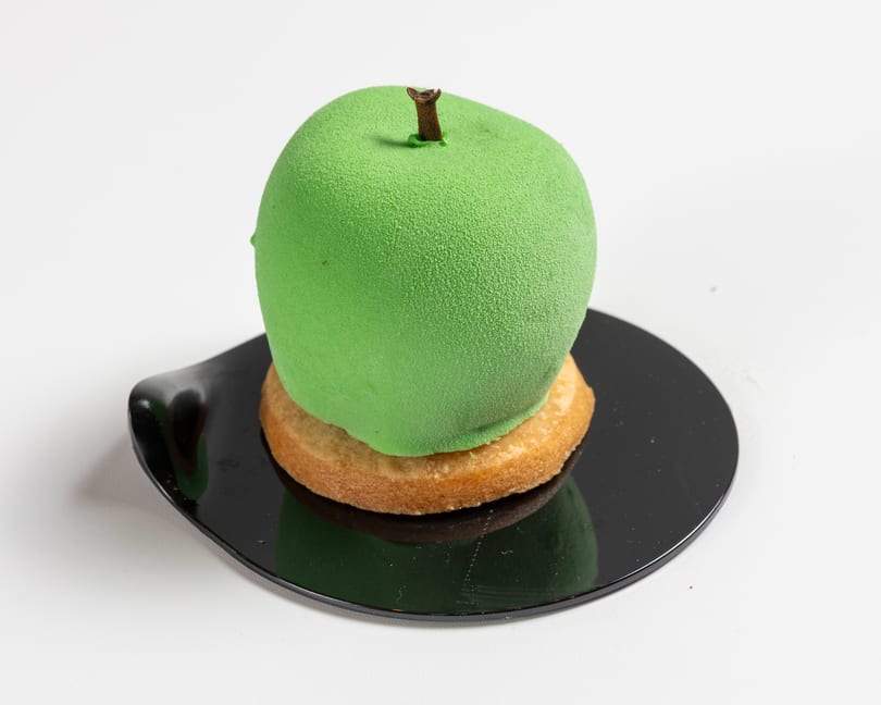 Green apple-shaped dessert with a brown stem on a round cookie and black plate.