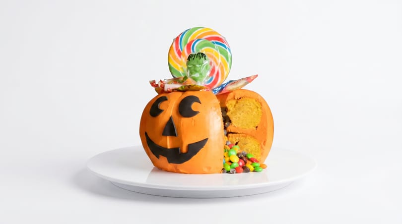 A Halloween pumpkin cake, cut open to reveal candy and cake inside, with a lollipop on top.