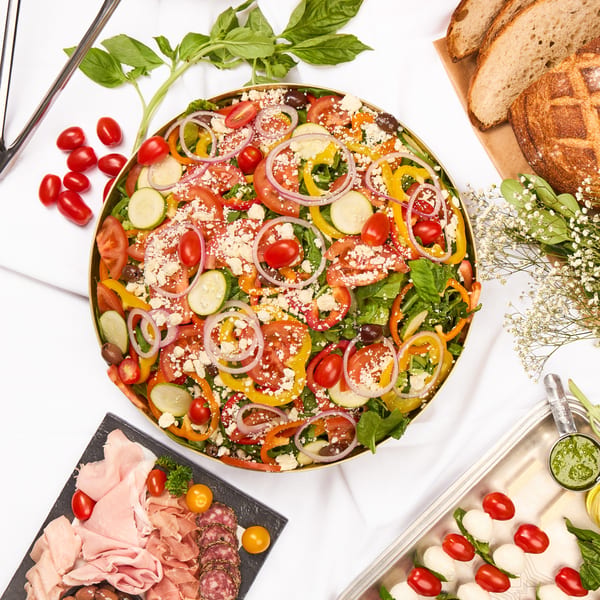 Overhead view of a buffet spread with salads, charcuterie, bread, and caprese skewers on a white tablecloth.