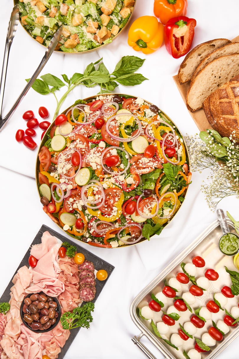 Overhead view of a buffet spread with salads, charcuterie, bread, and caprese skewers on a white tablecloth.
