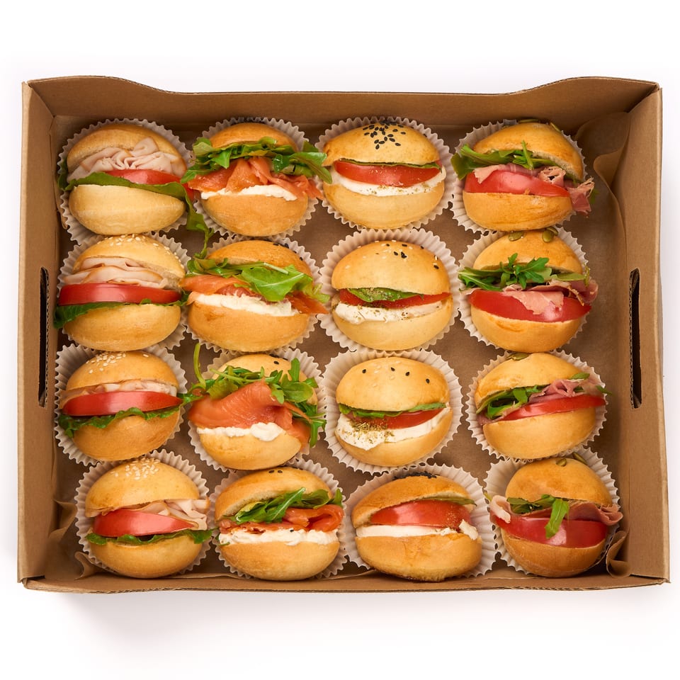 An overhead view of a cardboard box filled with 16 mini sandwiches, each in a white paper cup.