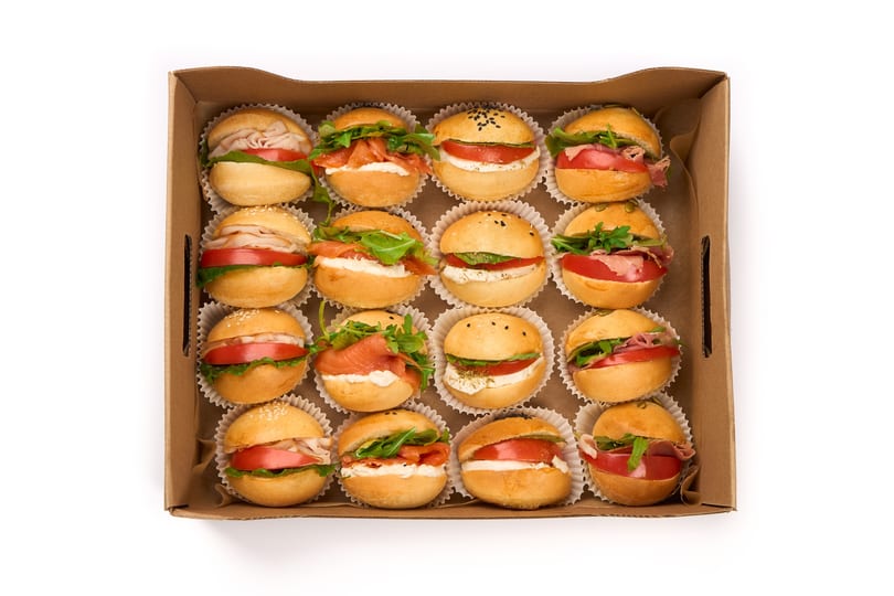 An overhead view of a cardboard box filled with 16 mini sandwiches, each in a white paper cup.