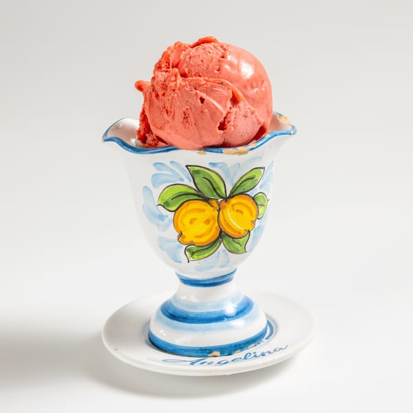 Scoop of pink ice cream in a decorative white cup with blue and yellow lemon design.