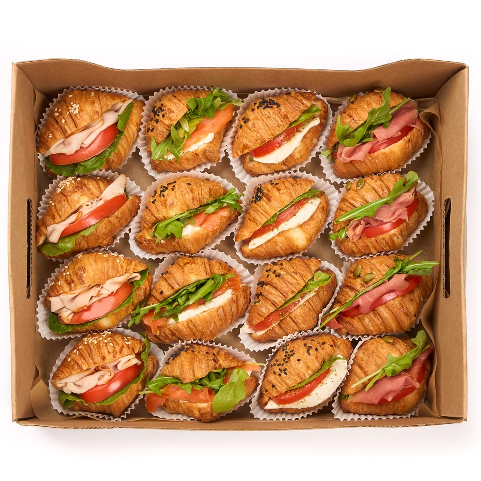 A cardboard box filled with 15 mini croissant sandwiches, each with various fillings.