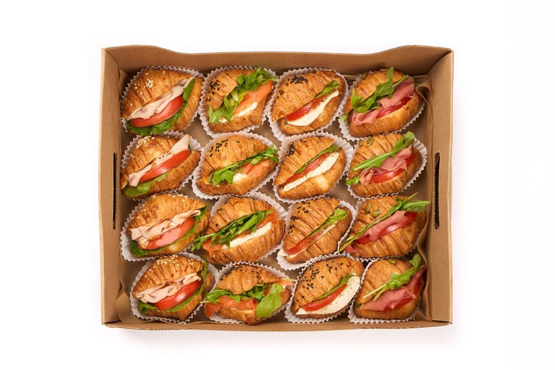 A cardboard box filled with 15 mini croissant sandwiches, each with various fillings.