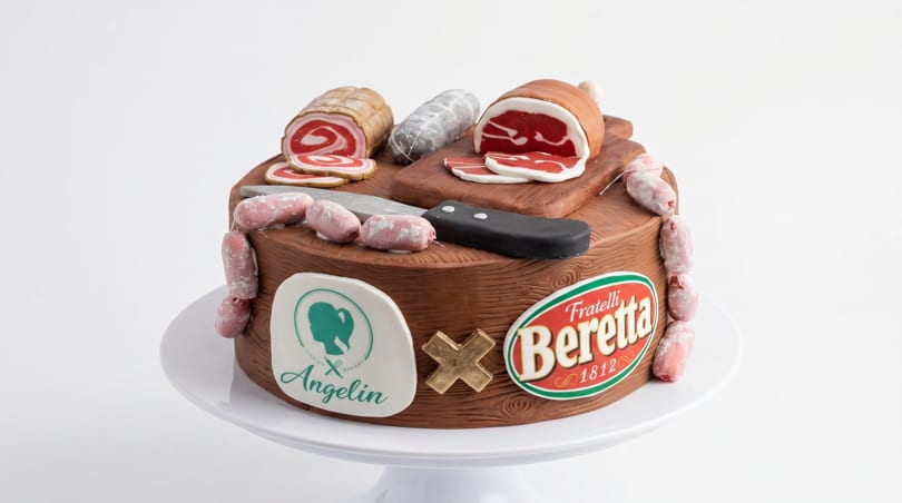 A cake designed to look like a charcuterie board with various meats, a knife, and brand logos.