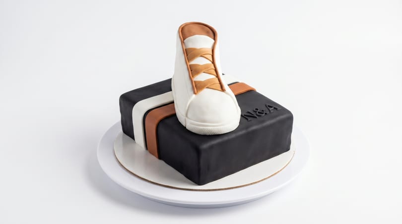 A black square cake with white and brown stripes, topped with a white and brown sneaker.