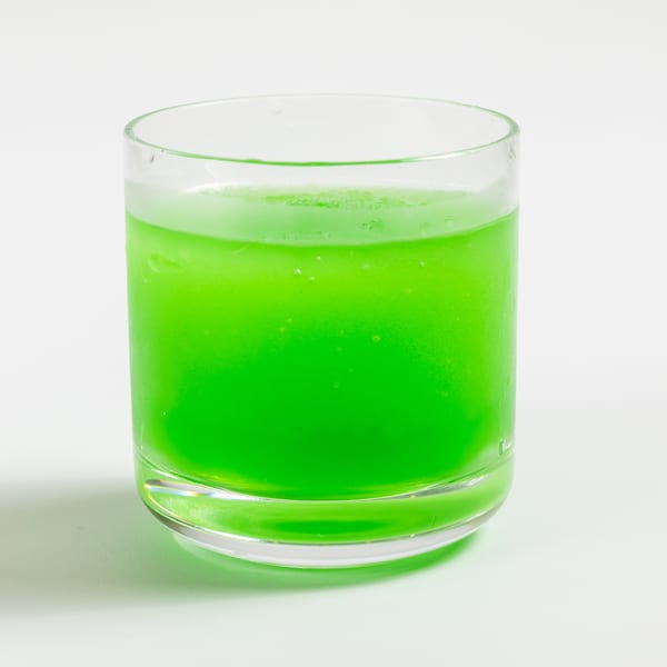 A vibrant green drink in a clear glass, with condensation and bubbles, on a white background.