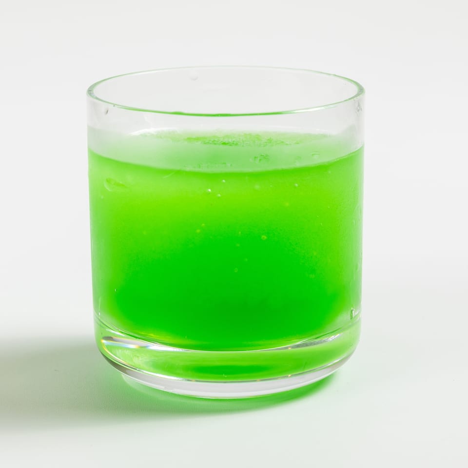 A vibrant green drink in a clear glass, with condensation and bubbles, on a white background.
