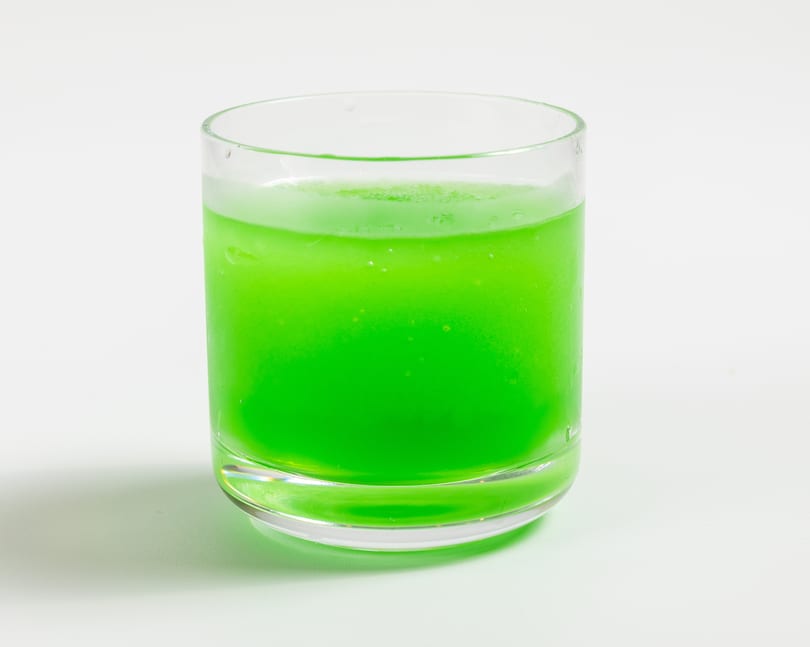 A vibrant green drink in a clear glass, with condensation and bubbles, on a white background.