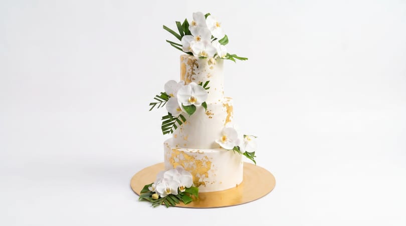 A three-tiered white wedding cake with gold leaf detailing, white orchids, and tropical greenery on a gold base.