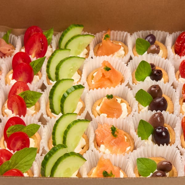 A box of assorted mini tarts with prosciutto, cucumber, salmon, olives, and tomatoes.