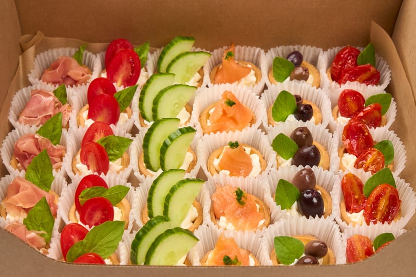 A box of assorted mini tarts with prosciutto, cucumber, salmon, olives, and tomatoes.