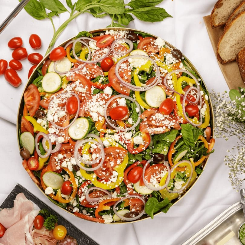 Large colorful salad with tomatoes, peppers, onions, zucchini, and feta cheese, surrounded by bread and charcuterie.