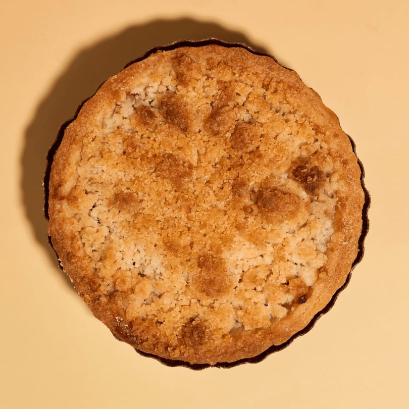 Overhead view of a golden-brown crumble tart with a scalloped edge on a light yellow surface.