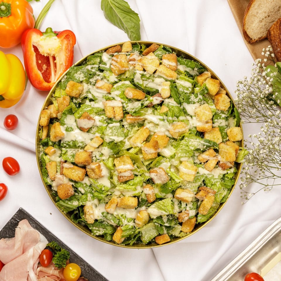 A large golden bowl of Caesar salad with croutons and dressing, surrounded by fresh vegetables.