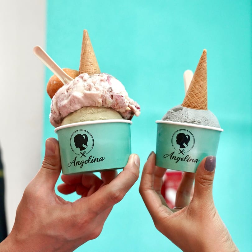 Two hands hold Angelina gelato cups with ice cream, waffle cones, and a light blue background.