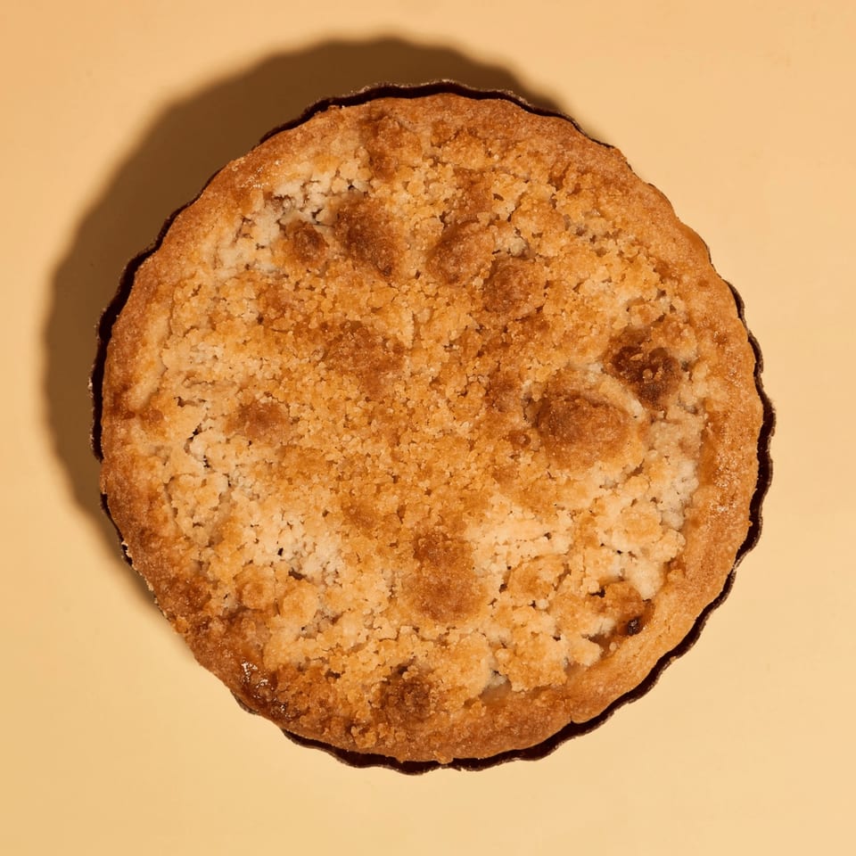 Overhead view of a golden-brown crumble tart with a scalloped edge on a light yellow surface.