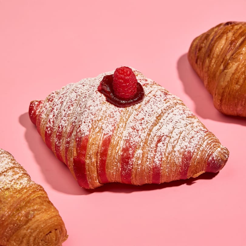 Raspberry croissant with powdered sugar and a fresh raspberry on a pink background.