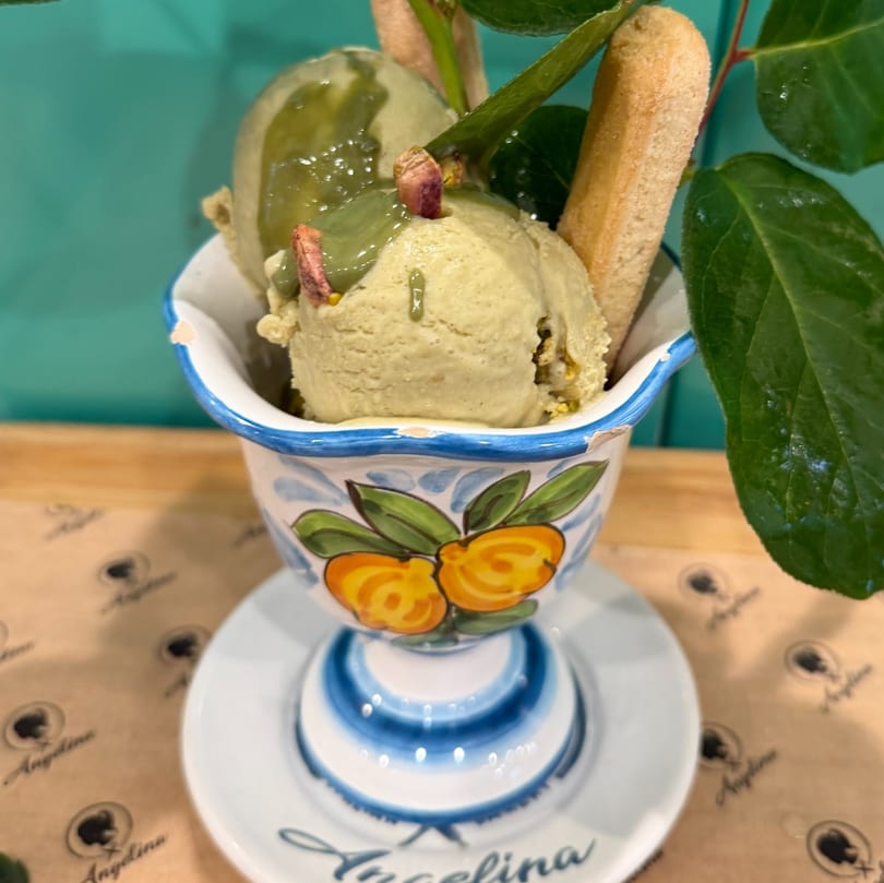 Pistachio gelato with ladyfingers and green leaves in a decorative ceramic bowl on a branded coaster.