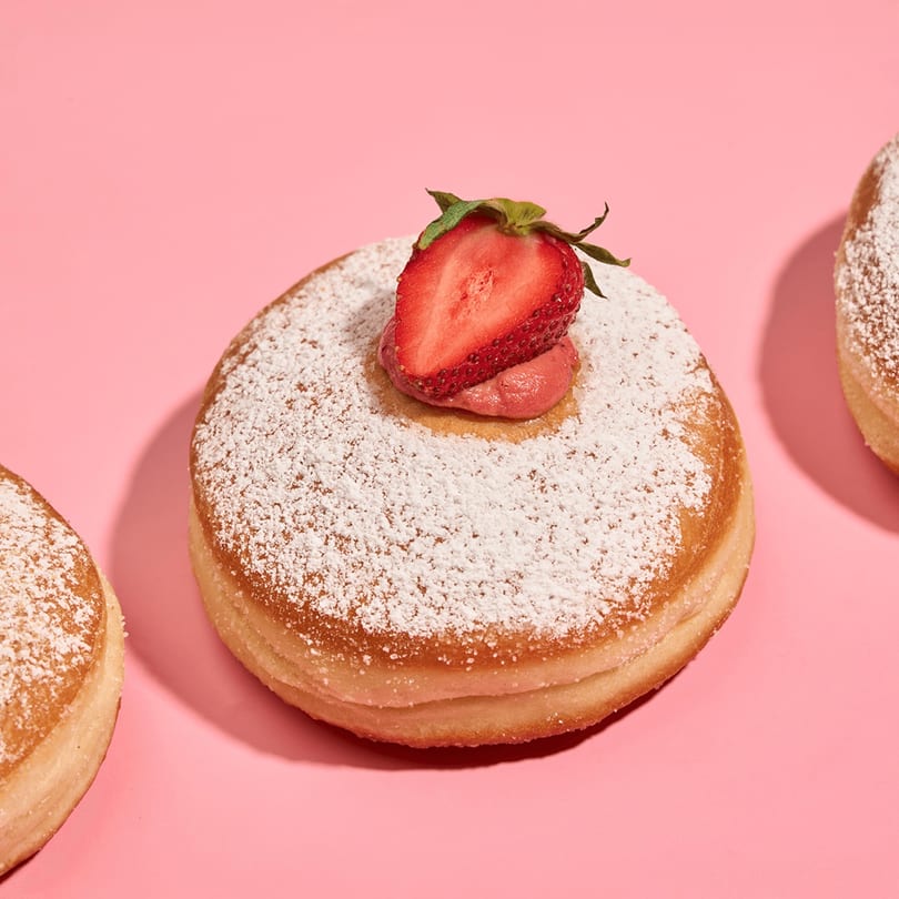 Powdered sugar donut with strawberry cream and a fresh strawberry half on a pink background.