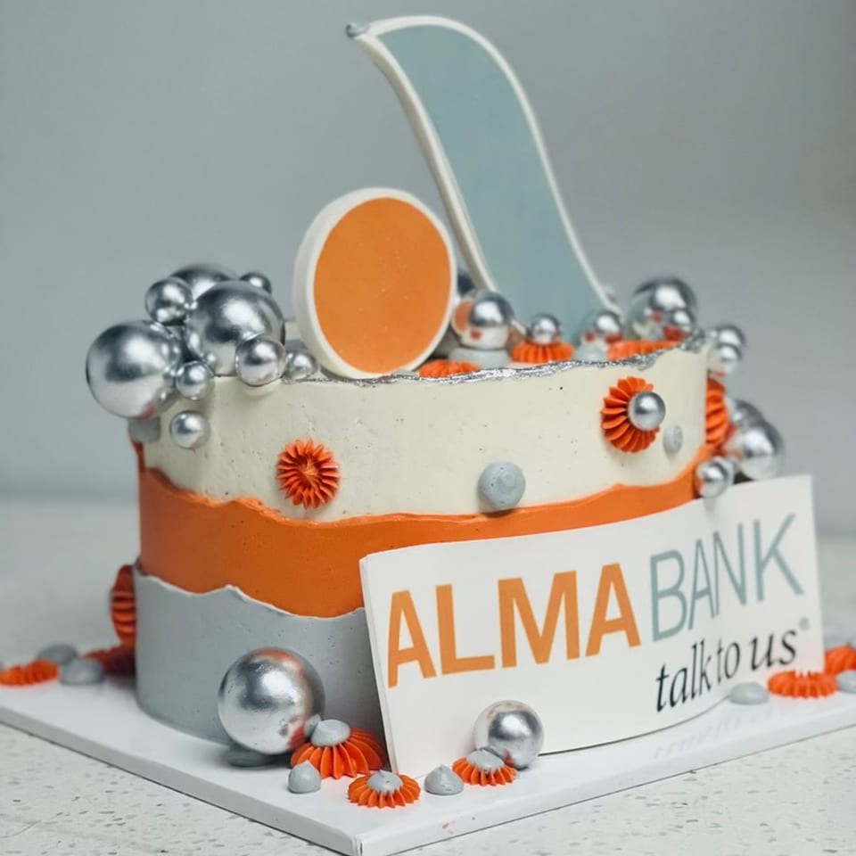A two-tier cake with orange, white, and gray frosting, silver spheres, and an "ALMA BANK" sign.