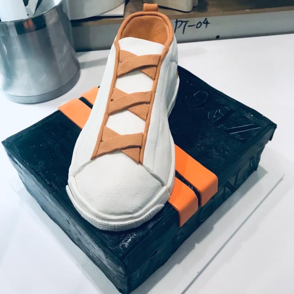 A white sneaker cake with orange laces sits atop a black rectangular cake with orange stripes.