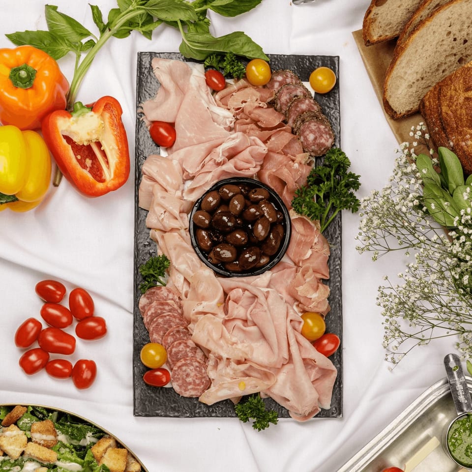 A charcuterie board with various meats, olives, cherry tomatoes, and bell peppers on a white tablecloth.