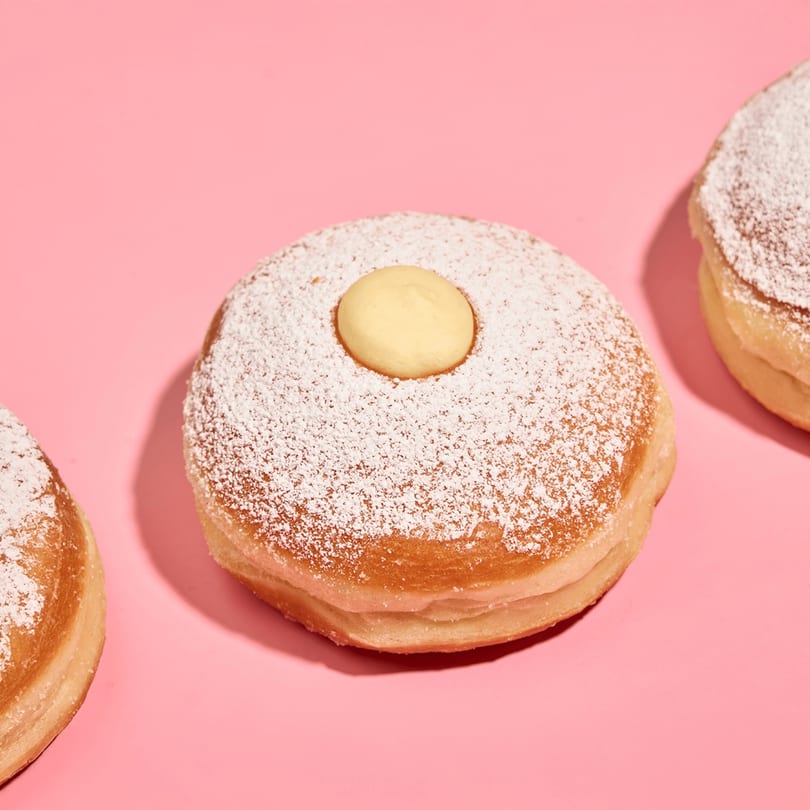 Three powdered sugar donuts with a dollop of cream in the center, on a pink background.