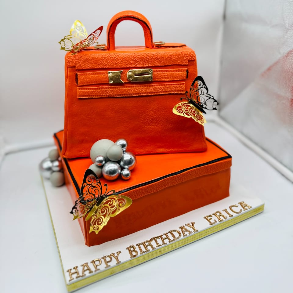 An orange handbag-shaped cake with gold butterflies and silver spheres, on a "Happy Birthday Erica" base.
