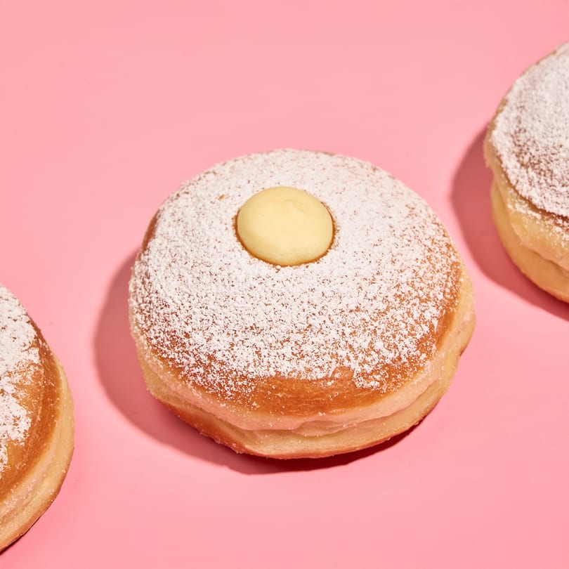 Three powdered sugar donuts with a dollop of cream in the center, on a pink background.