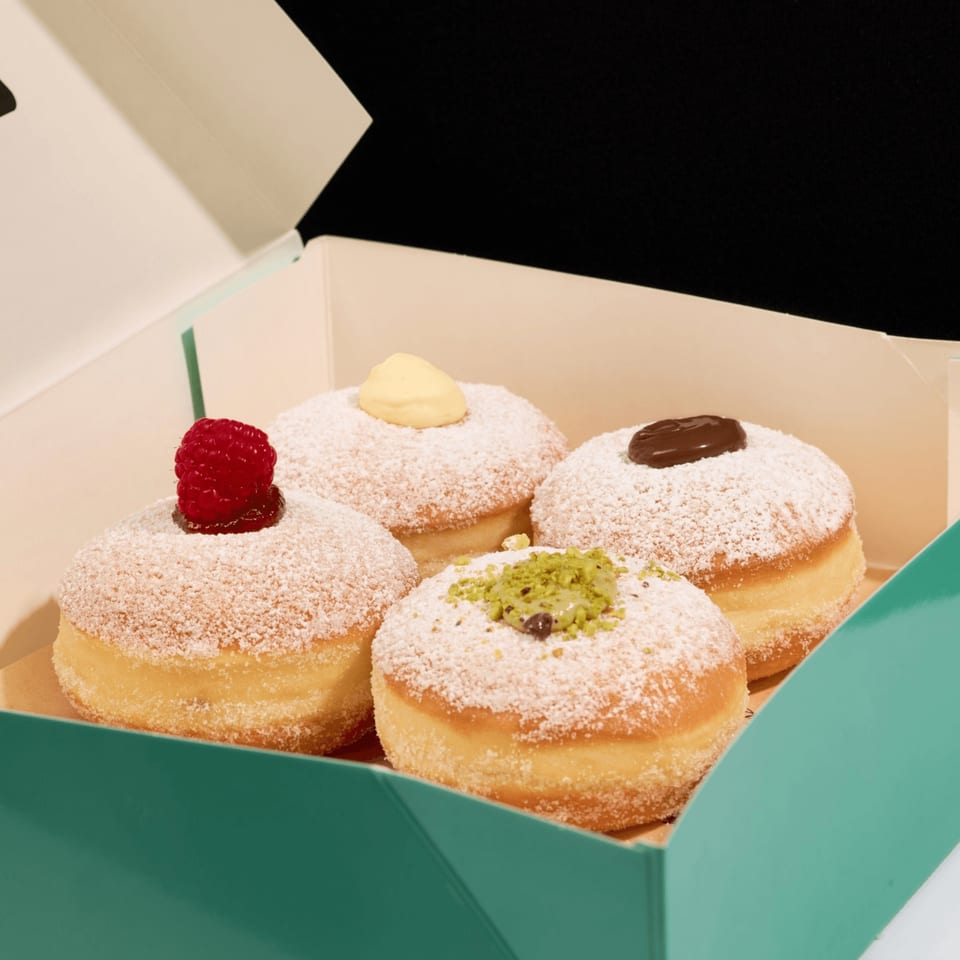 Four powdered sugar donuts with raspberry, vanilla, chocolate, and pistachio toppings in a box.