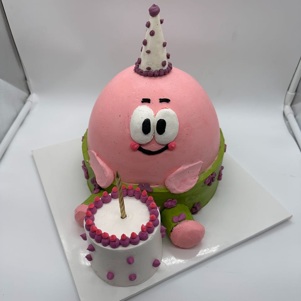 A pink Patrick Star cake wearing a party hat, sitting next to a small white birthday cake with a candle.