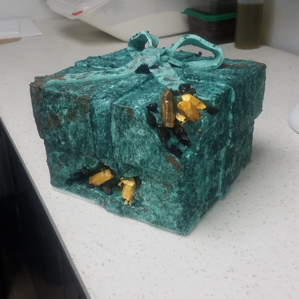 A teal gift box cake with a bow, adorned with black and gold edible gems.