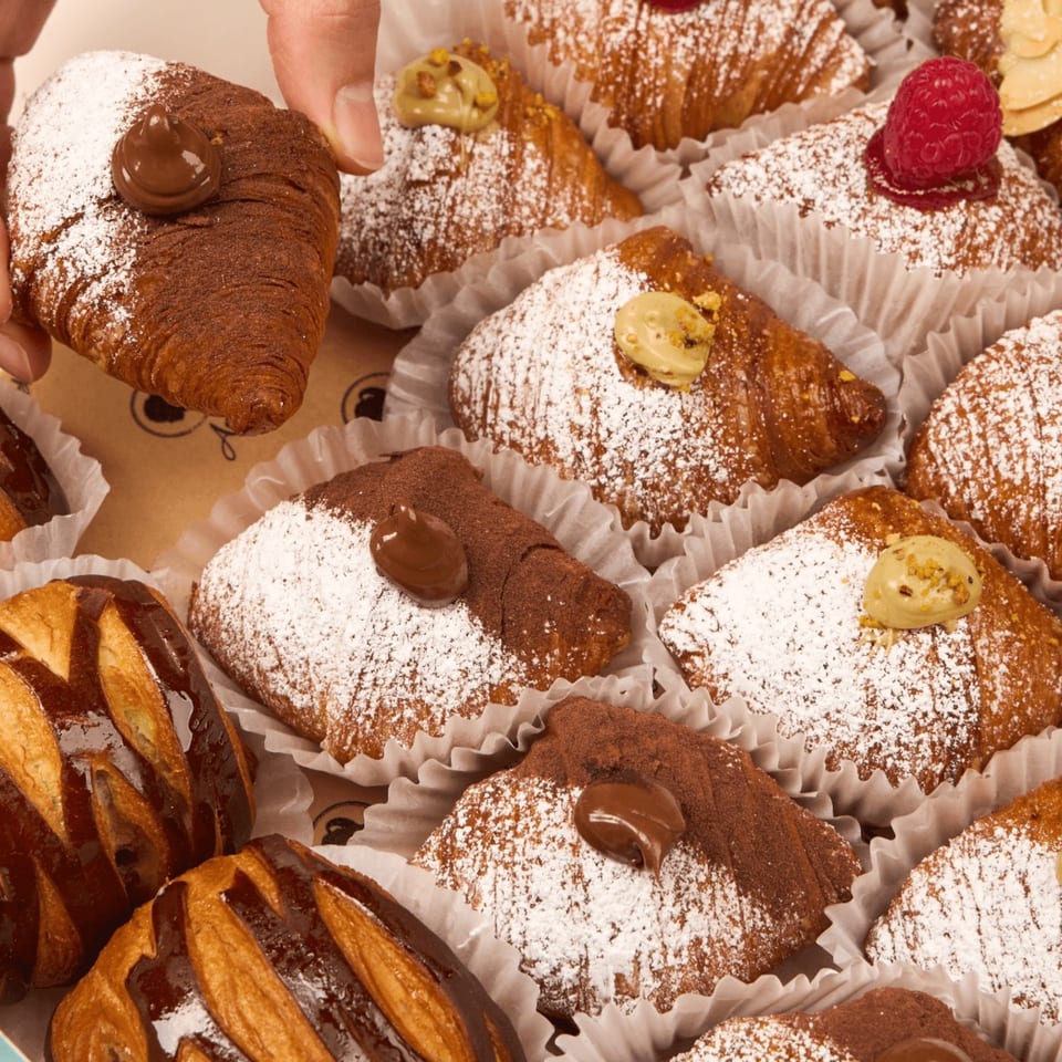 A hand picks up a chocolate croissant from a box of assorted pastries dusted with powdered sugar.