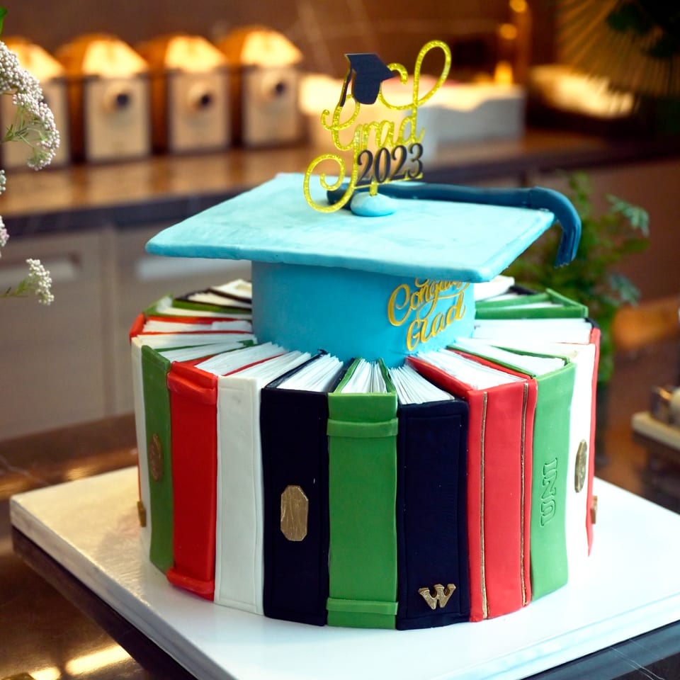 A graduation cake shaped like a blue cap and a stack of colorful books with a "Grad 2023" topper.
